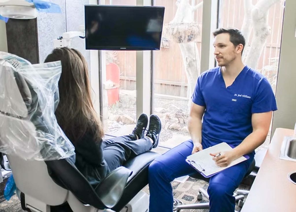 Dentist in blue scrubs talking with a patient sitting in a dental chair.